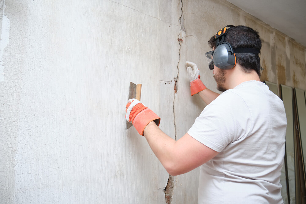 Ouvrier du bâtiment réparant une fissure et appliquant du ciment sur un mur. L'ouvrier porte des gants de sécurité, des lunettes et un casque antibruit.
