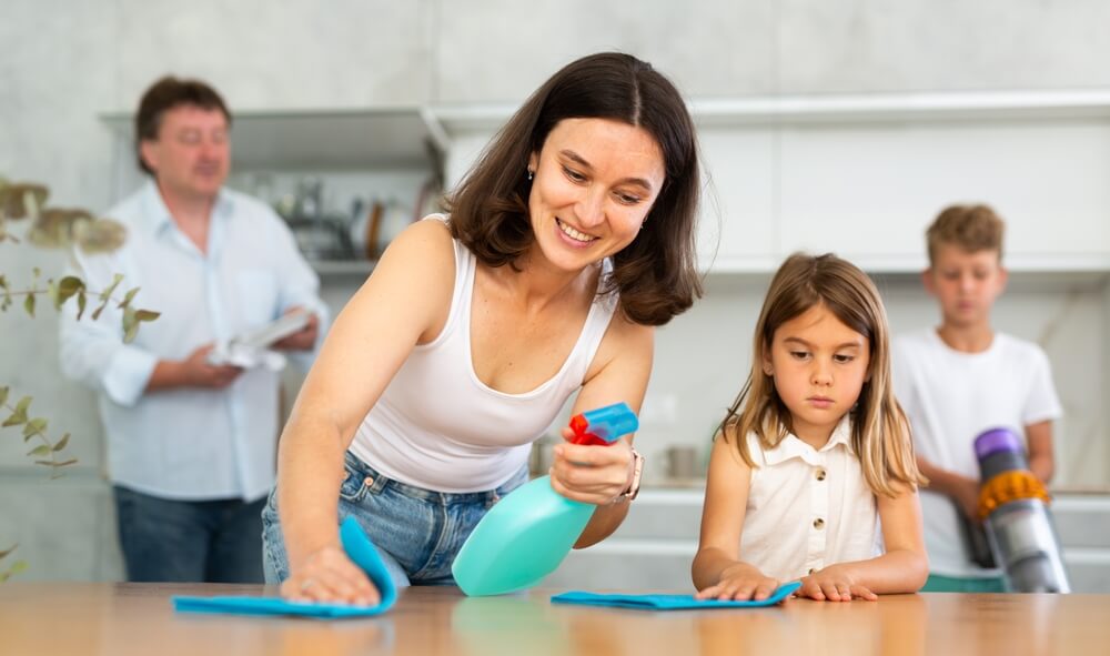 Femme positive lave la table de cuisine avec chiffon avec sa fille. Mari et fils aident à faire le nettoyage dans la cuisine en arrière-plan. Nettoyage de maison familiale, tâches ménagères, aide ménagère.